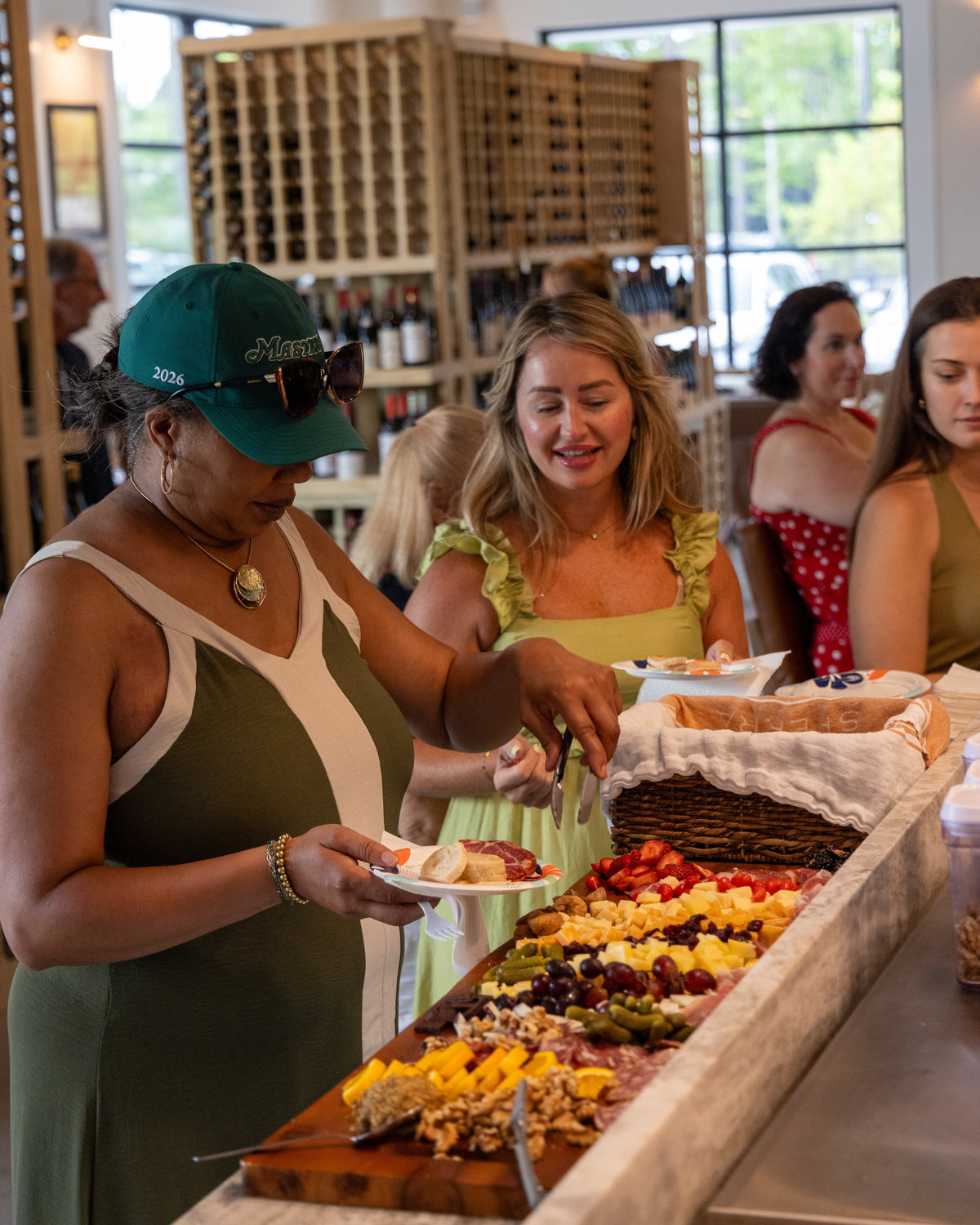 A woman in a green cap and olive dress serves food from a large charcuterie board lined up in a bright indoor event space with wine racks in the background.
