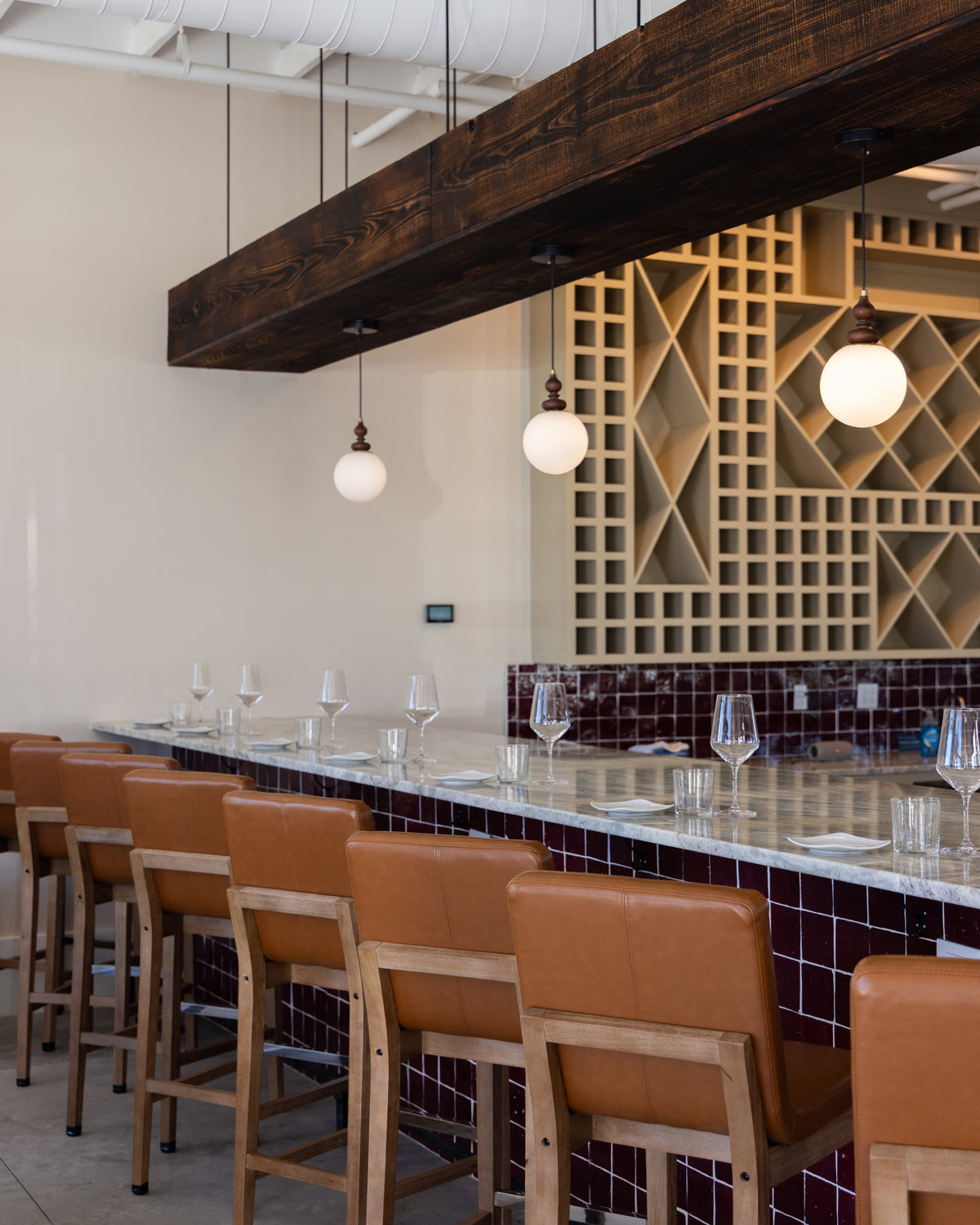 A modern bar interior with a long counter, brown leather and wood bar stools, hanging globe pendant lights, a dark wood ceiling beam, and a geometric wood wall backdrop.