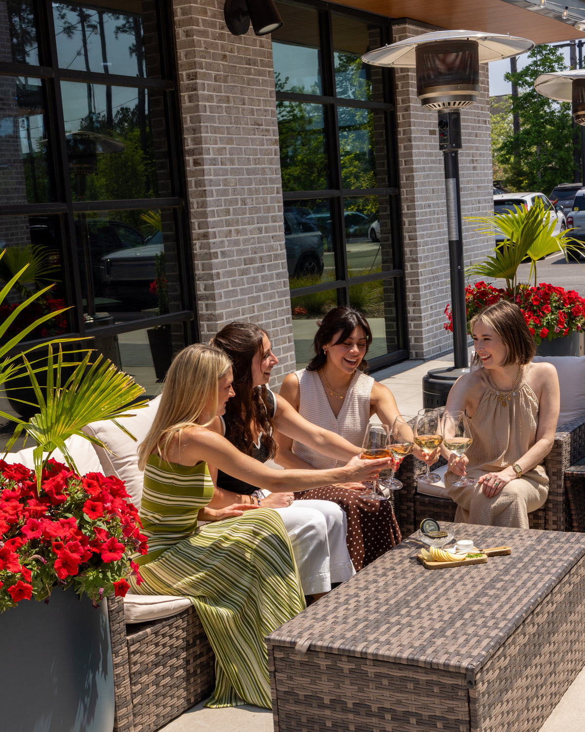 Four women laughing and toasting with wine glasses on an outdoor patio, seated on woven wicker furniture next to a coffee table, with brick columns, large windows, and potted red flowers in the background.