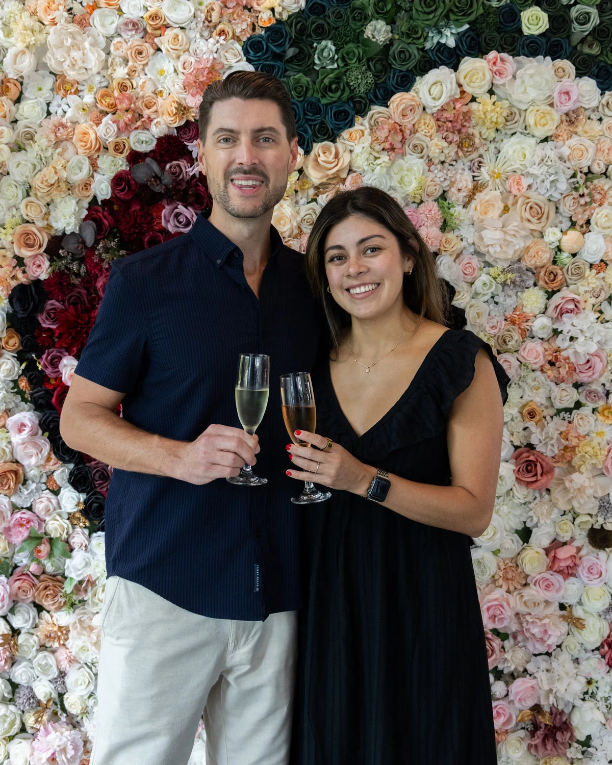A smiling man and woman standing together in front of a large floral wall backdrop, each holding a champagne flute. The man wears a dark navy short-sleeve button-up shirt and light beige pants, while the woman wears a black sleeveless dress.